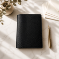 A black textured leather planner or notebook cover lies closed on a light marble surface, styled with a gold pen, folded white paper, and a small potted plant in soft diagonal sunlight.